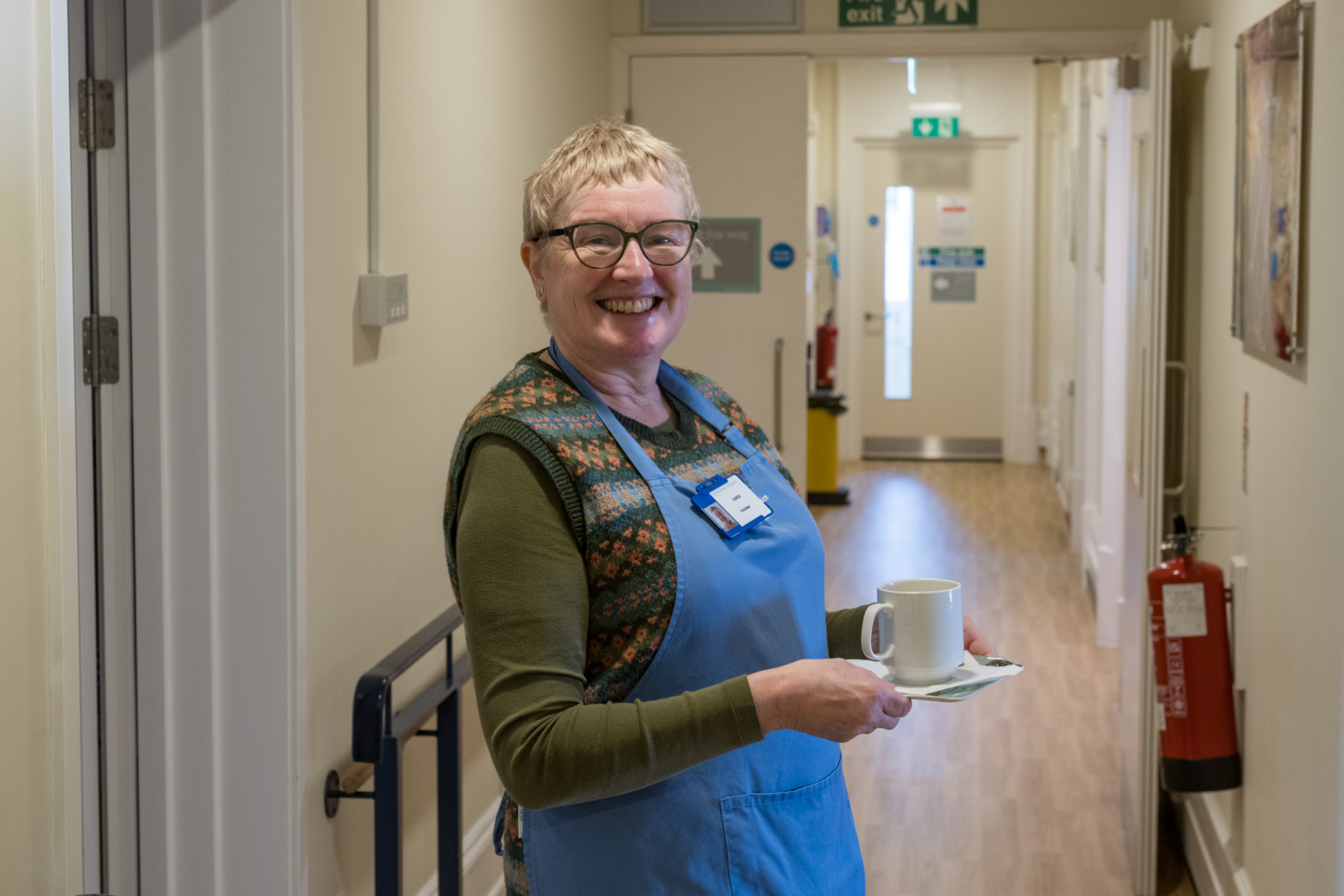 A ward volunteer carrying a cup of tea smiling at the camera